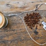 Top-down view of a latte with foam art, a pile of coffee beans, and white earphones on a weathered wooden table.