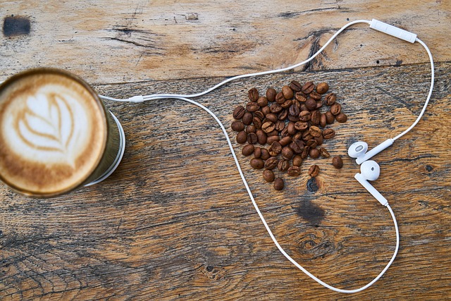 Top-down view of a latte with foam art, a pile of coffee beans, and white earphones on a weathered wooden table.