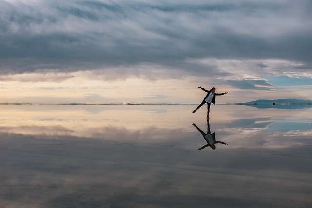 A figure stands arms outstretched and on one foot on a beach, in shallow water, with the reflection clearly visible. In indie publishing, the journey is never over. You just move on to the next stage.